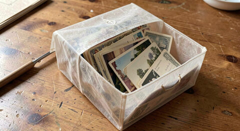 A glassine sleeve box filled with sorted old postcards standing on a plain wooden table